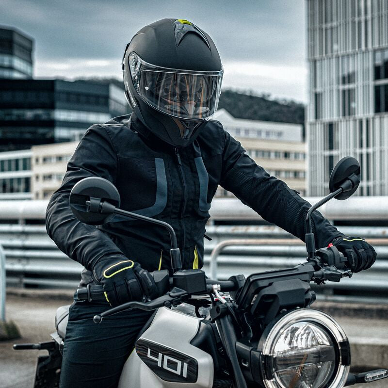 A person wearing a black full-face motorcycle helmet, black leather riding jacket, gloves, and pants sits on a black and white motorcycle on an urban bridge with modern buildings in the background.