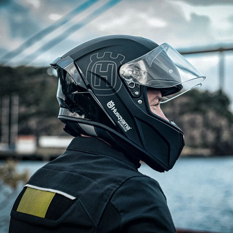 A side-profile view of a rider wearing a matte black Husqvarna full-face motorcycle helmet with a clear visor, set against a scenic background of water and a bridge.