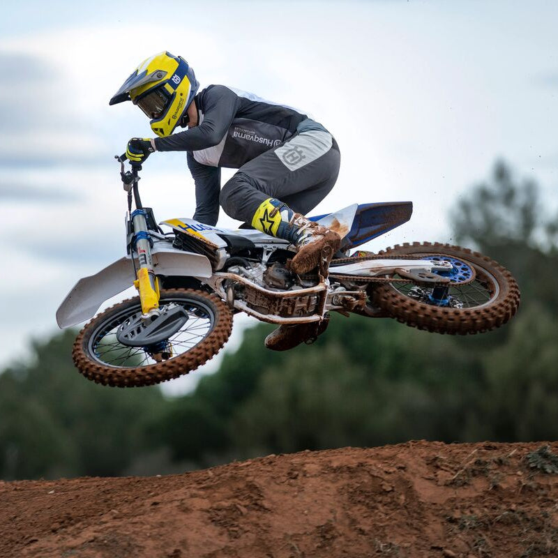 A motocross rider in full gear jumps a dirt bike mid-air over a dirt track, with trees and an overcast sky in the background.
