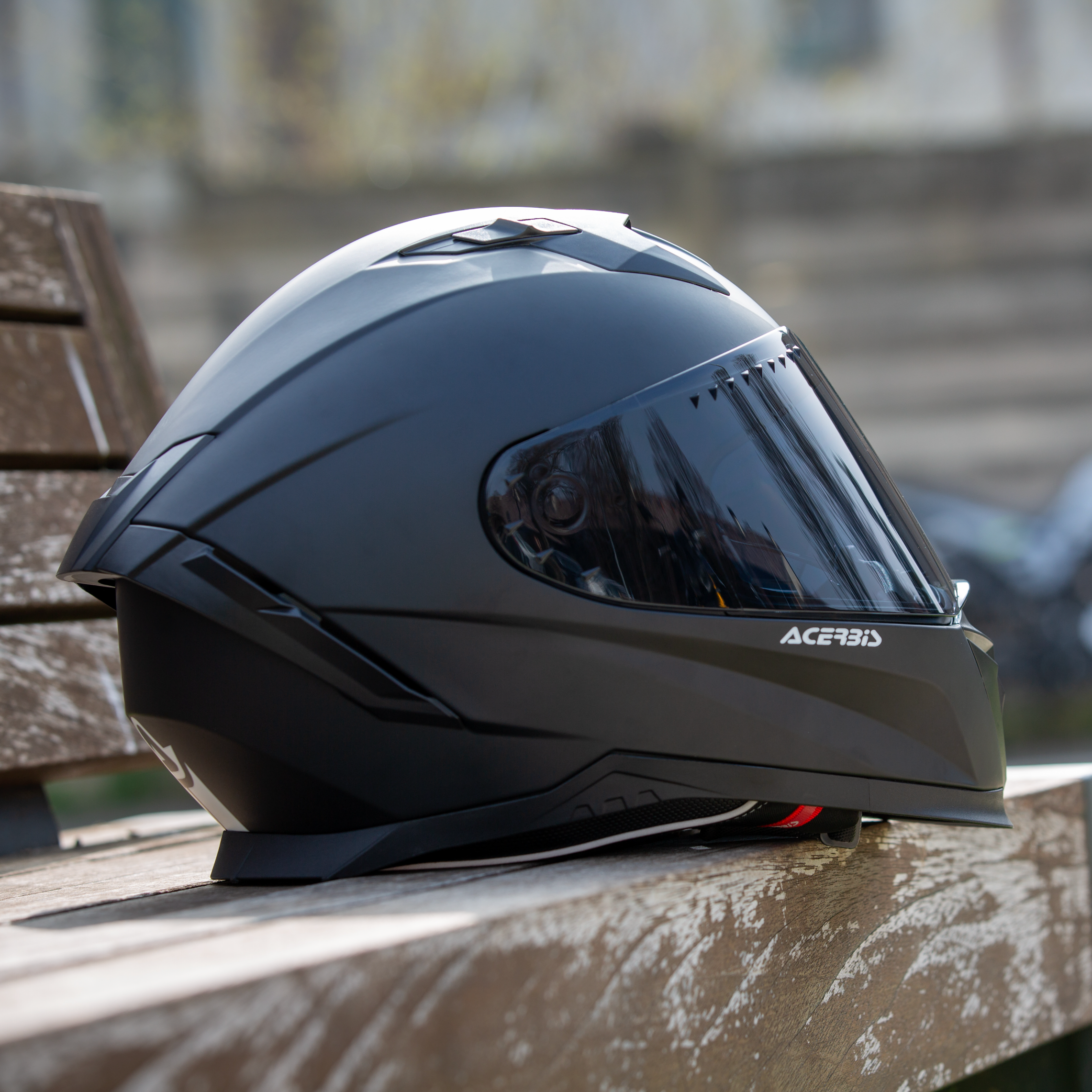 Black Acerbis motorcycle helmet on a wooden bench with a blurred motorcycle in the background.