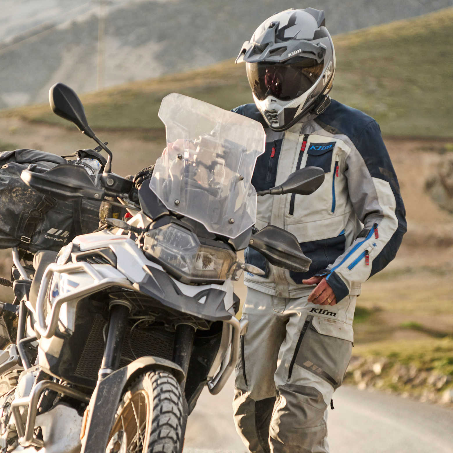 Motorcyclist in protective gear walking beside a motorcycle on a dirt road with mountains in the background