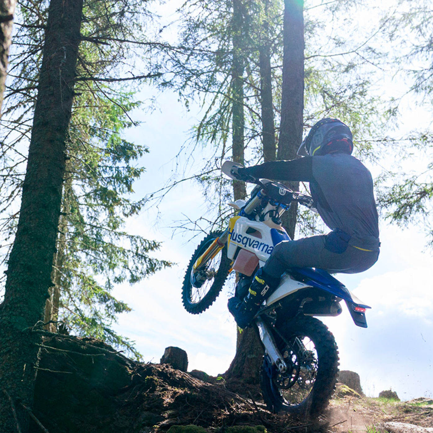 A rider wearing a helmet and protective gear maneuvers a blue and white Husqvarna dirt bike up a steep, rocky forest trail, with tall pine trees and bright sunlight filtering through the canopy.