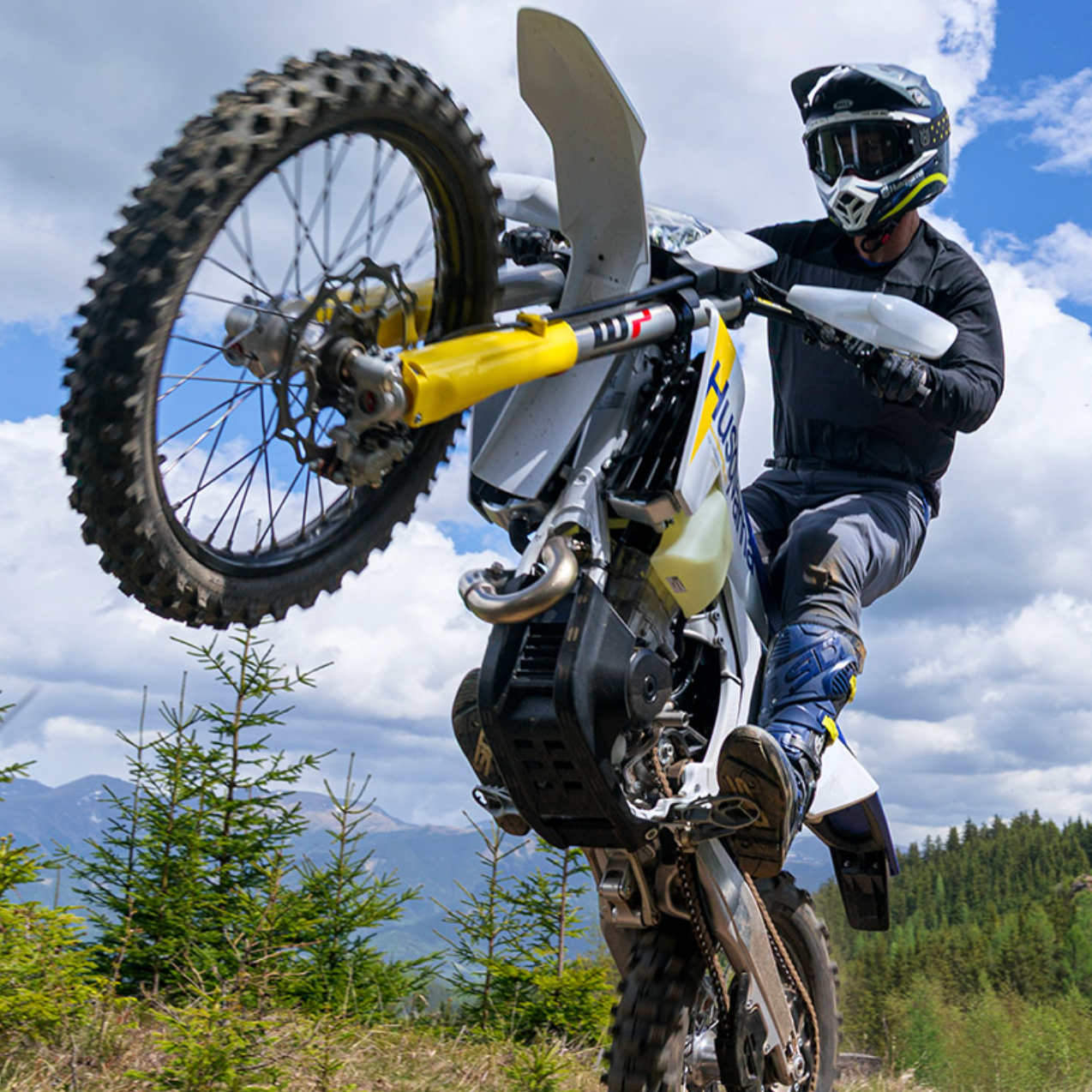 A person riding a white and yellow off-road motorcycle performing a wheelie outdoors on a grassy hill with trees and mountains in the background under a blue sky with clouds.