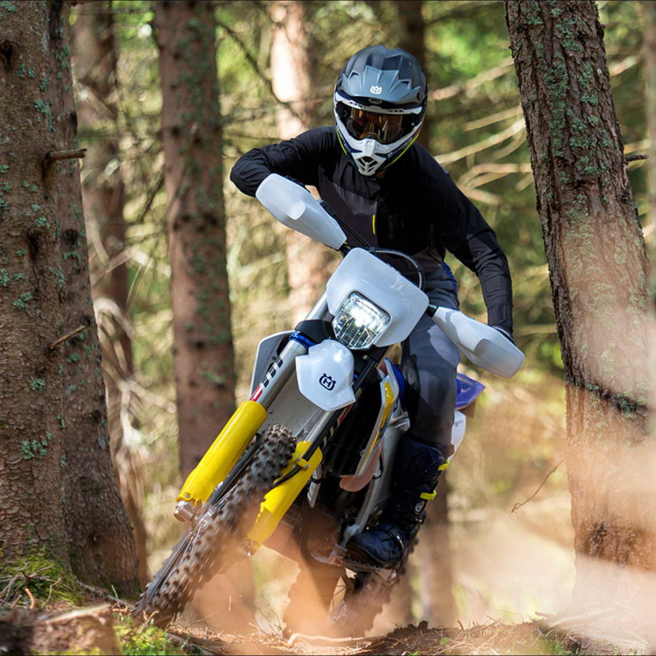 A person riding a white and yellow off-road motorcycle through a dense forest, wearing a black helmet, goggles, and protective gear.