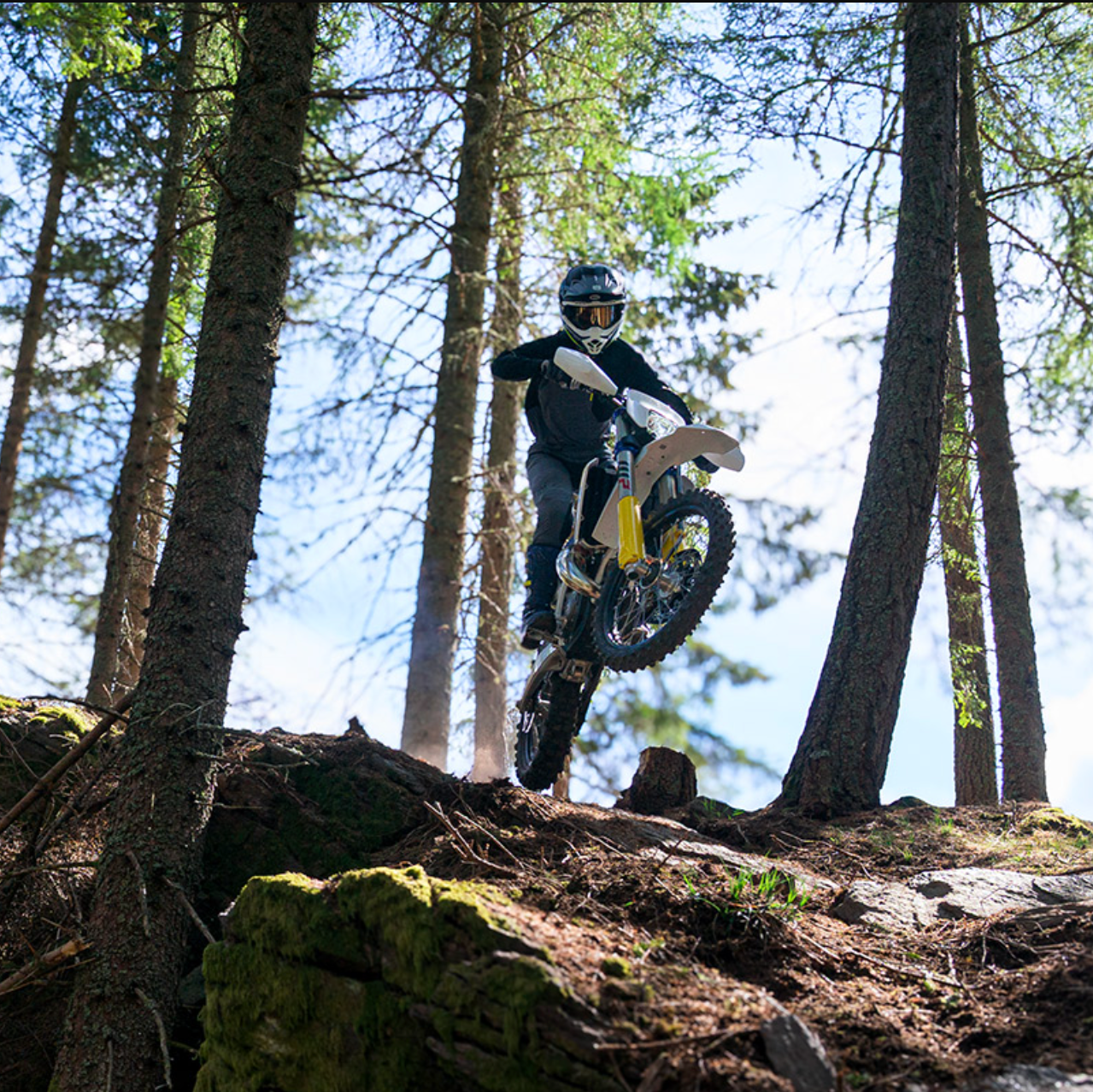 A person riding a dirt bike or motocross bike through a forest, with tall trees and a rocky, moss-covered trail visible. The rider is wearing full protective gear and the bike has a yellow accent.