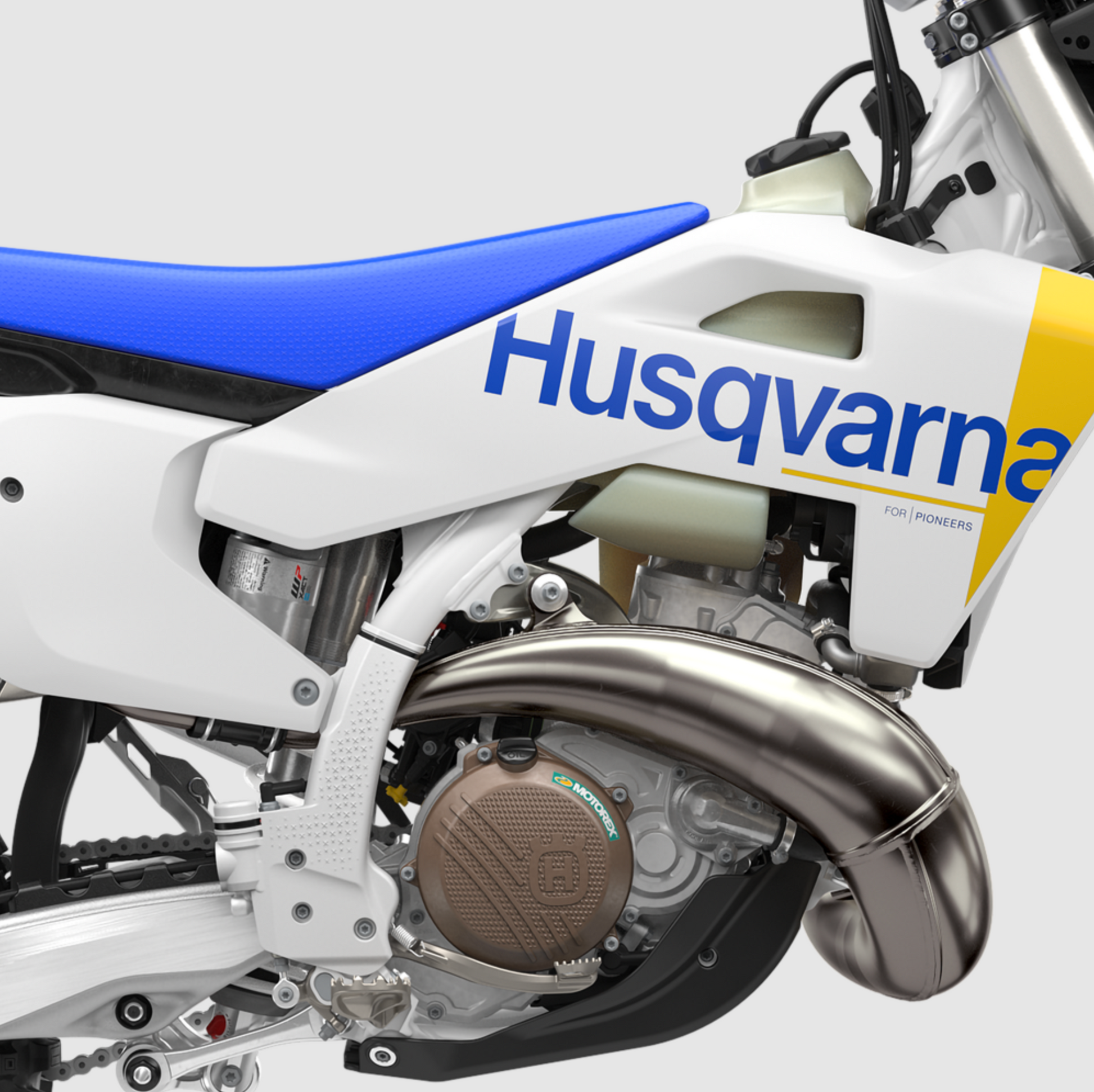 Close-up of a Husqvarna dirt bike showing the white bodywork with blue and yellow graphics, a blue seat, and a polished silver exhaust pipe against a light gray background.