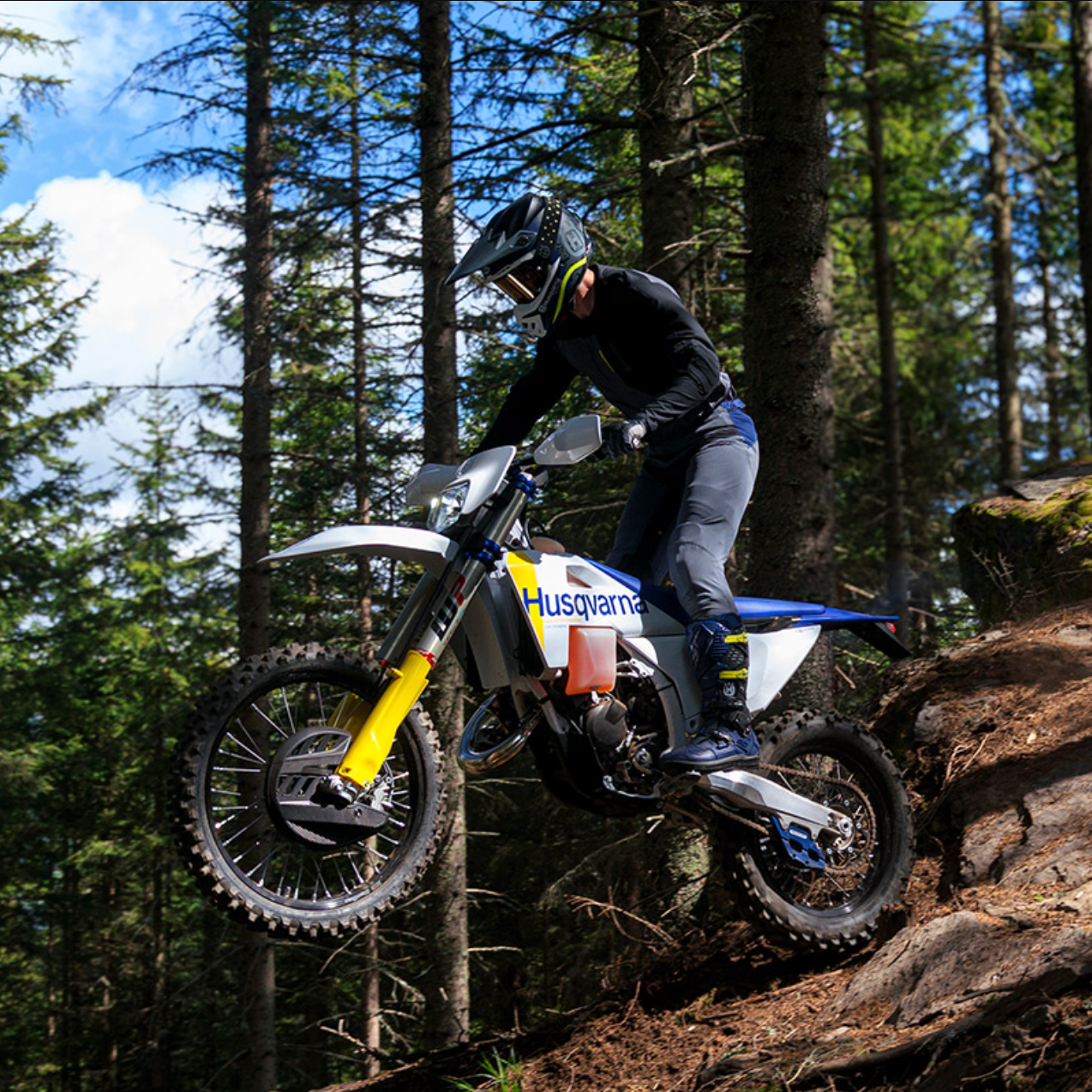 A rider wearing a black helmet and protective gear maneuvers a Husqvarna dirt bike up a steep, rocky forest trail surrounded by tall pine trees.