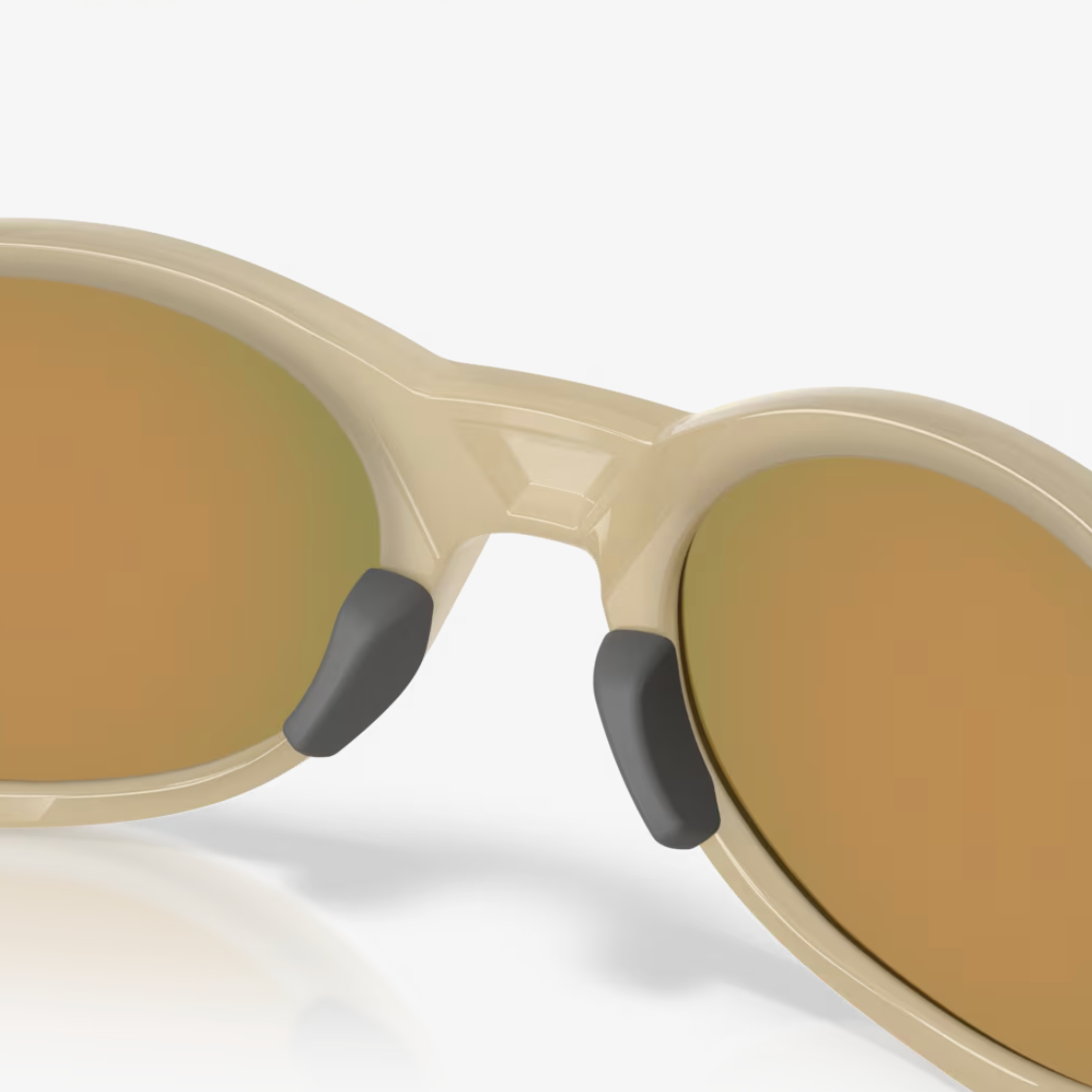 Close-up of oval sunglasses with a warm beige frame and mirrored lenses reflecting a gold-brown tone, shown on a light gray background.