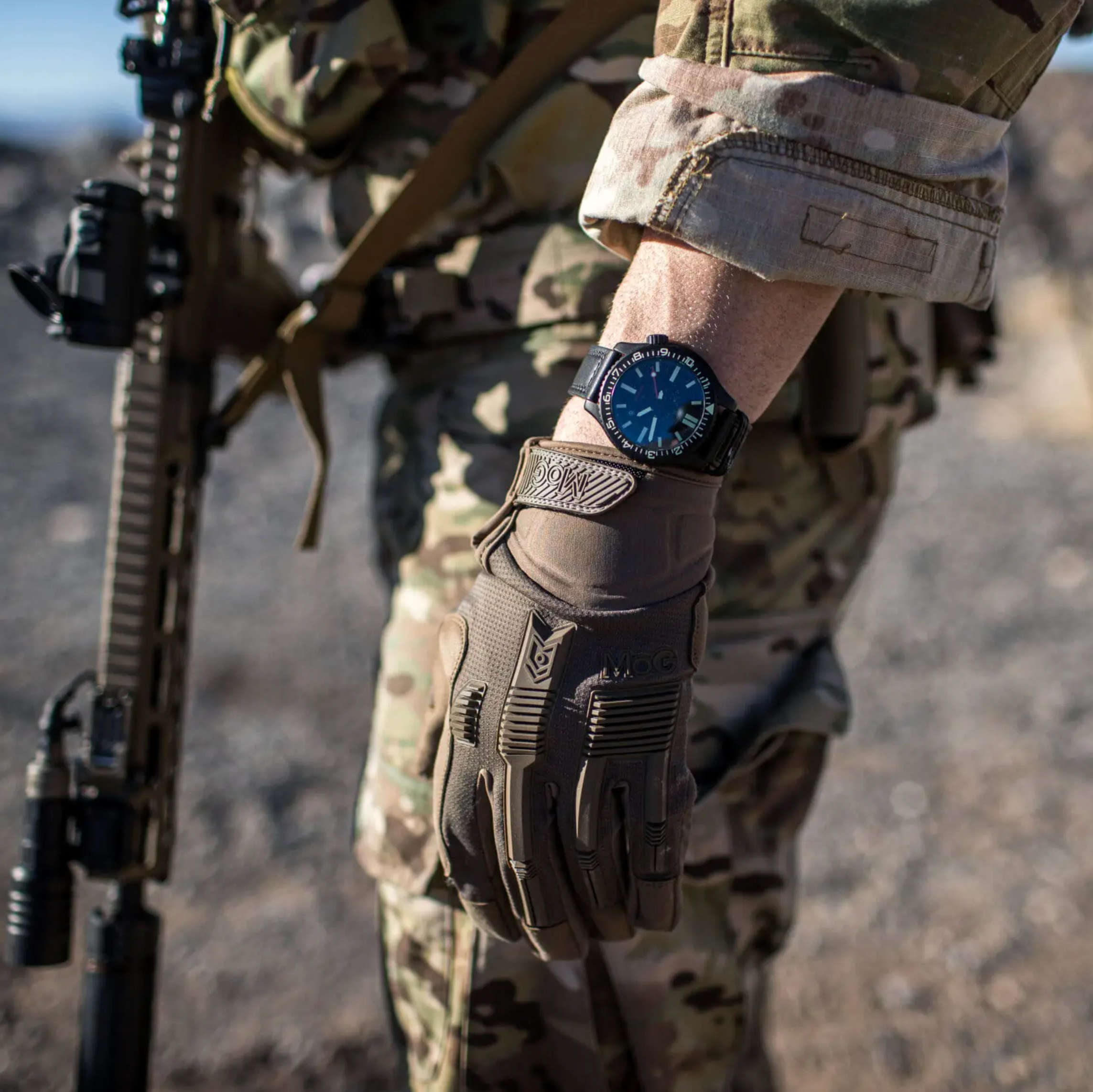 A close-up lifestyle shot of a person in camouflage military uniform wearing a tactical watch with a black strap and blue dial, holding a tactical glove and a rifle in an outdoor desert setting.