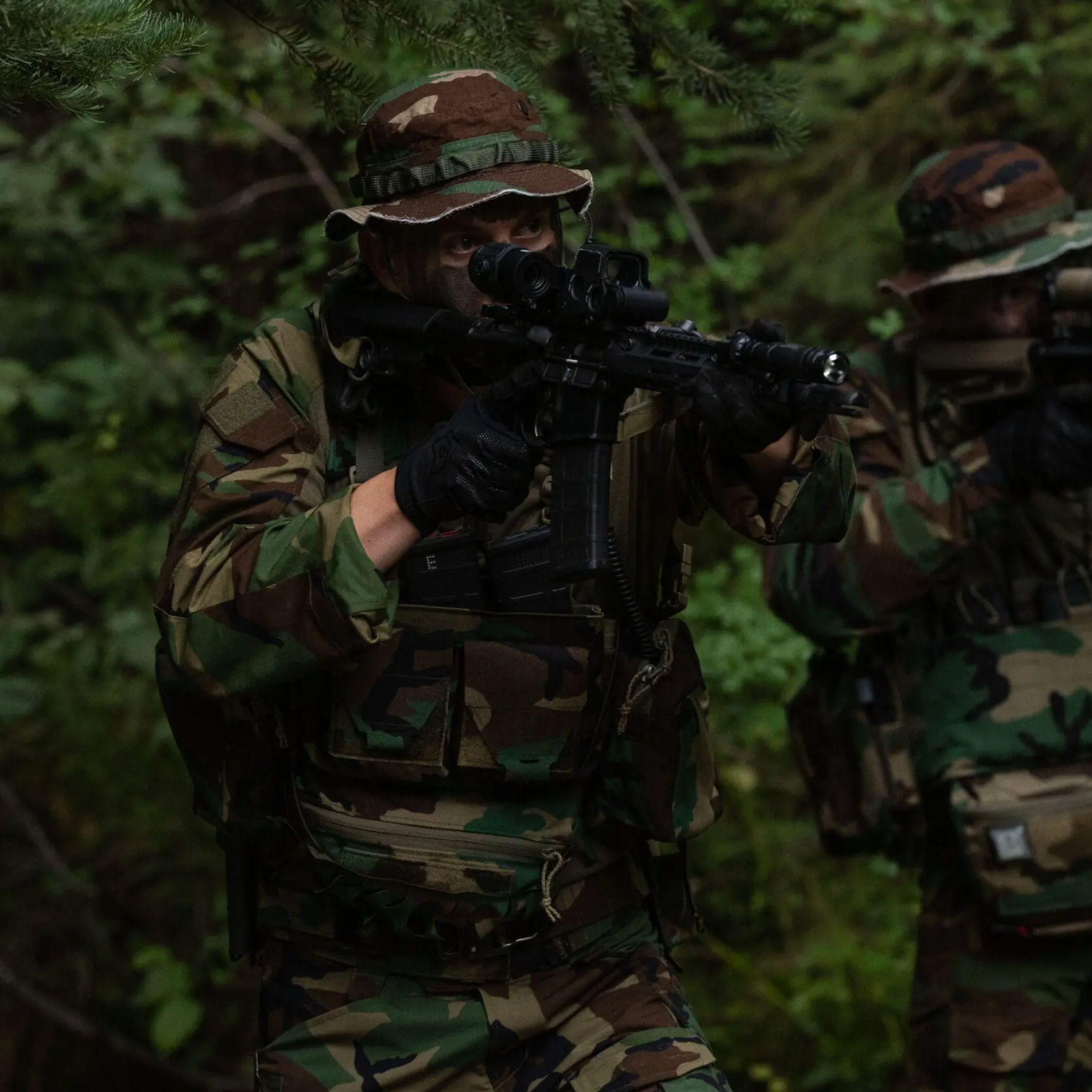 Two soldiers in woodland camouflage uniforms and bucket hats aiming rifles with scopes through dense green foliage.