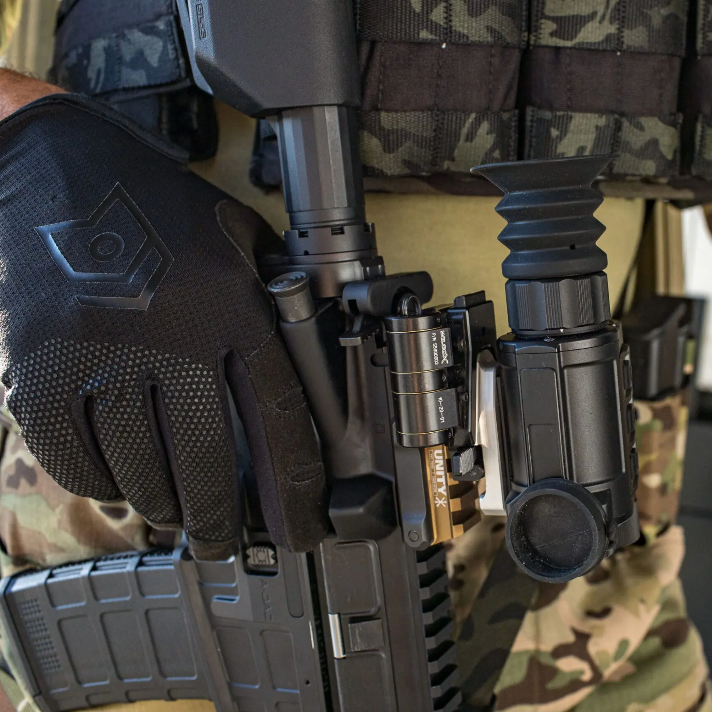Close-up of a person in camouflage tactical gear wearing a black textured glove and carrying a black rifle with mounted optic accessories.