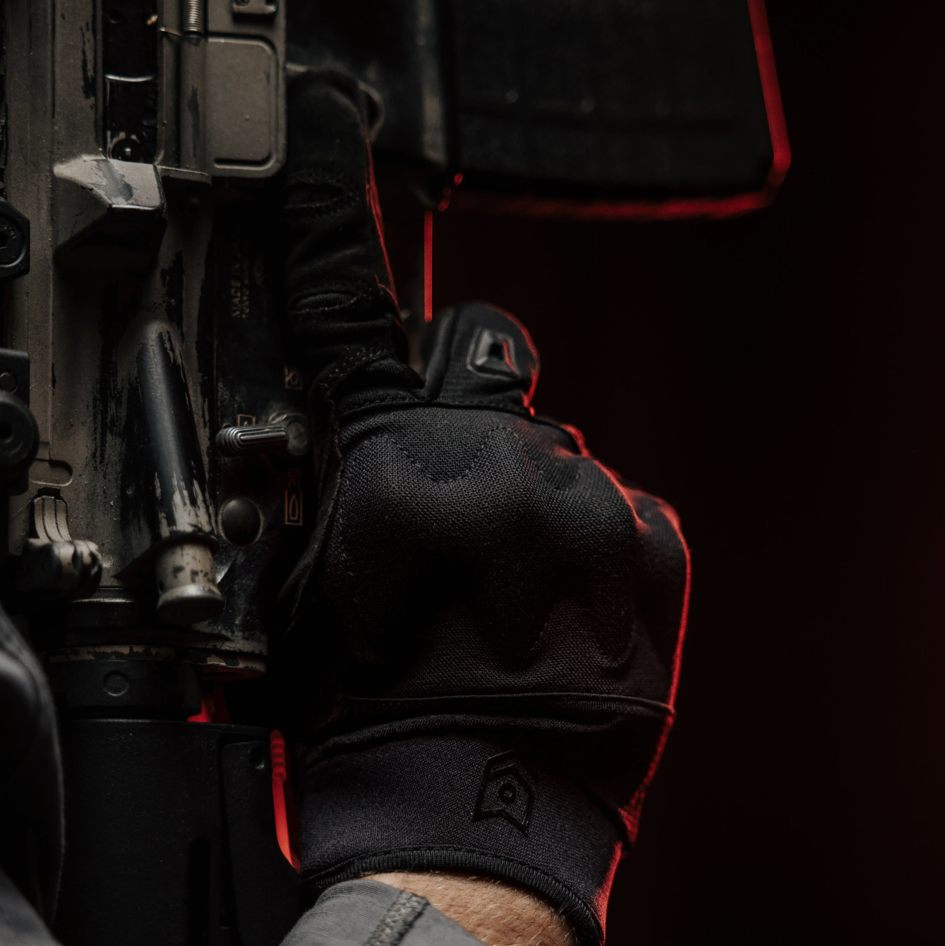 A close-up of a hand wearing a black tactical glove with red stitching, holding a firearm. The background is dark and shadowed, emphasizing the black and red color scheme of the glove.