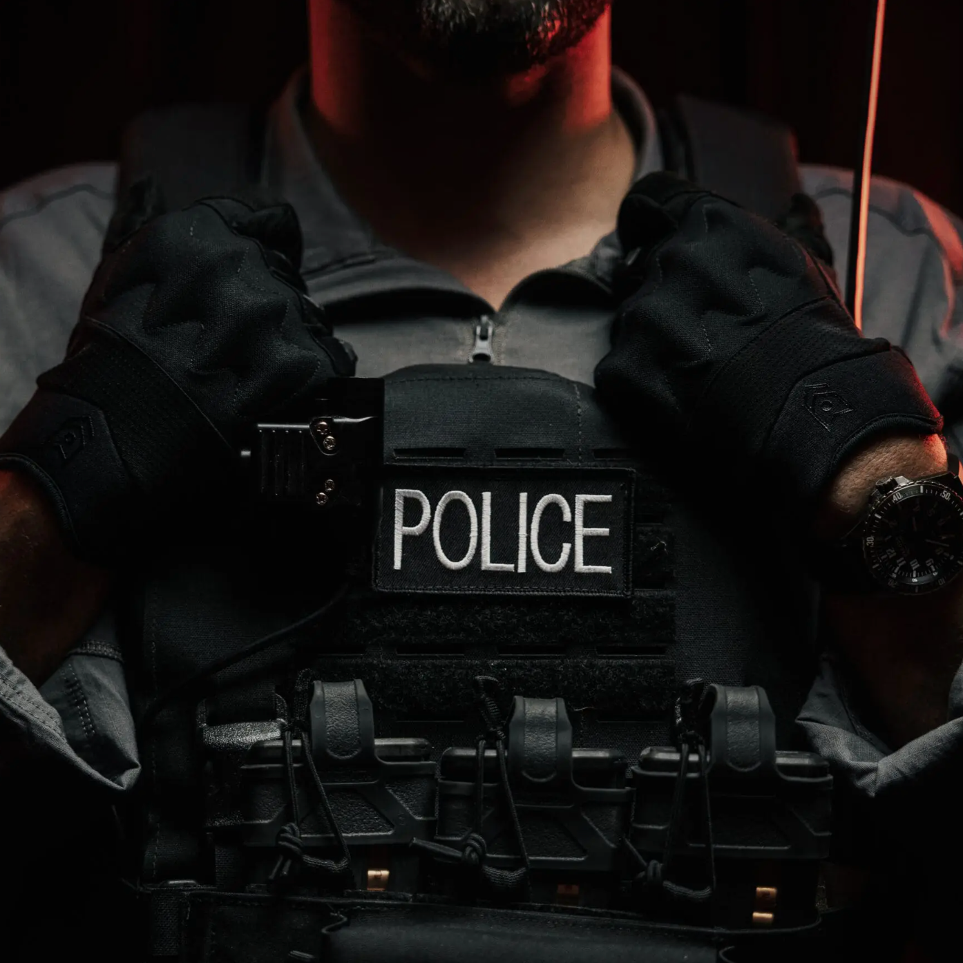 A close-up of a tactical police officer wearing a black vest with a white 'POLICE' name tape patch, black gloves, and a watch, set against a very dark background.