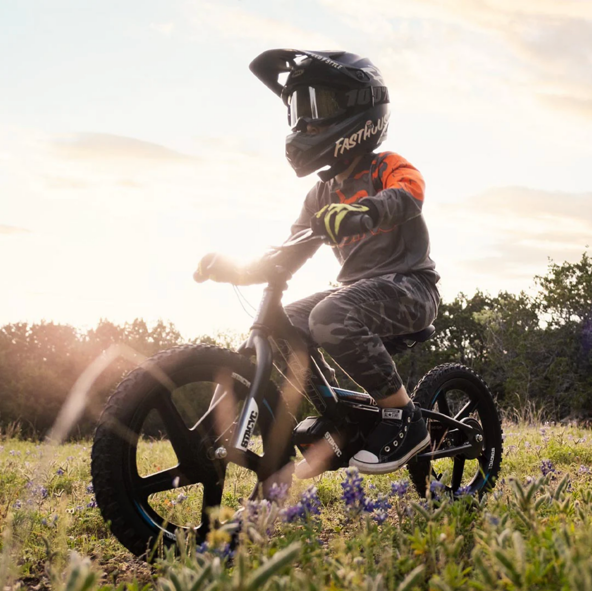 A child wearing a black full-face helmet and a gray long-sleeve jersey with orange accents rides a black balance bike on a grassy field at sunset. The helmet features the Fasttrack brand logo.